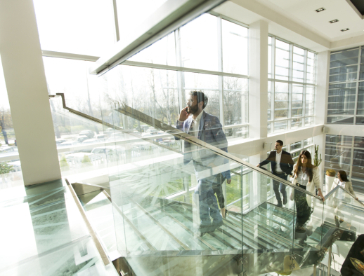 People walking up a glass staircase in a modern office building with large windows are immersed in engaging conversations. A man at the front, phone in hand, is discussing property line disputes as he ascends, blending work and movement seamlessly.