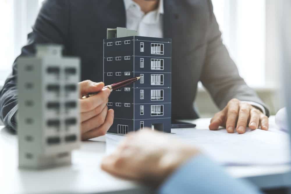 Two professionals engage in a detailed discussion over architectural models of buildings on a table, one thoughtfully pointing with a pencil, mindful of potential property line disputes.