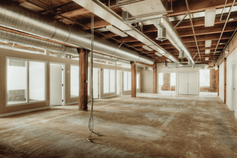 An empty industrial-style office space with exposed ductwork, wood beams, and unfinished floors awaits its new purpose. Glass-panel doors line one side of the room, offering a clear boundary that leaves no room for property line disputes within this urban oasis.
