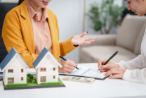 Two people sit at a table cluttered with model houses, documents, and cash. One gestures animatedly about property line disputes while the other diligently notes details on a clipboard.