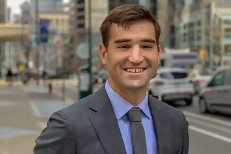 A man in a gray suit and tie is smiling outdoors on a city street with blurred traffic and buildings in the background.