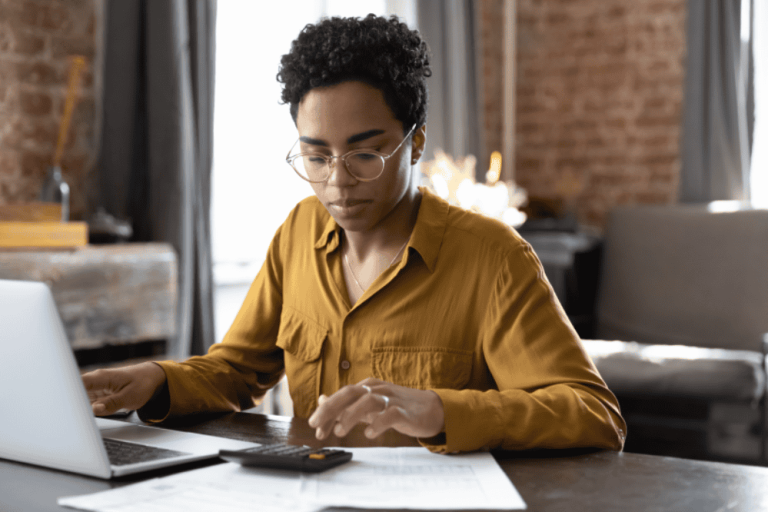 A person in glasses and a yellow shirt works diligently at a desk, navigating complex property line disputes with the aid of a laptop and calculator, all within the charming confines of a brick-walled room.