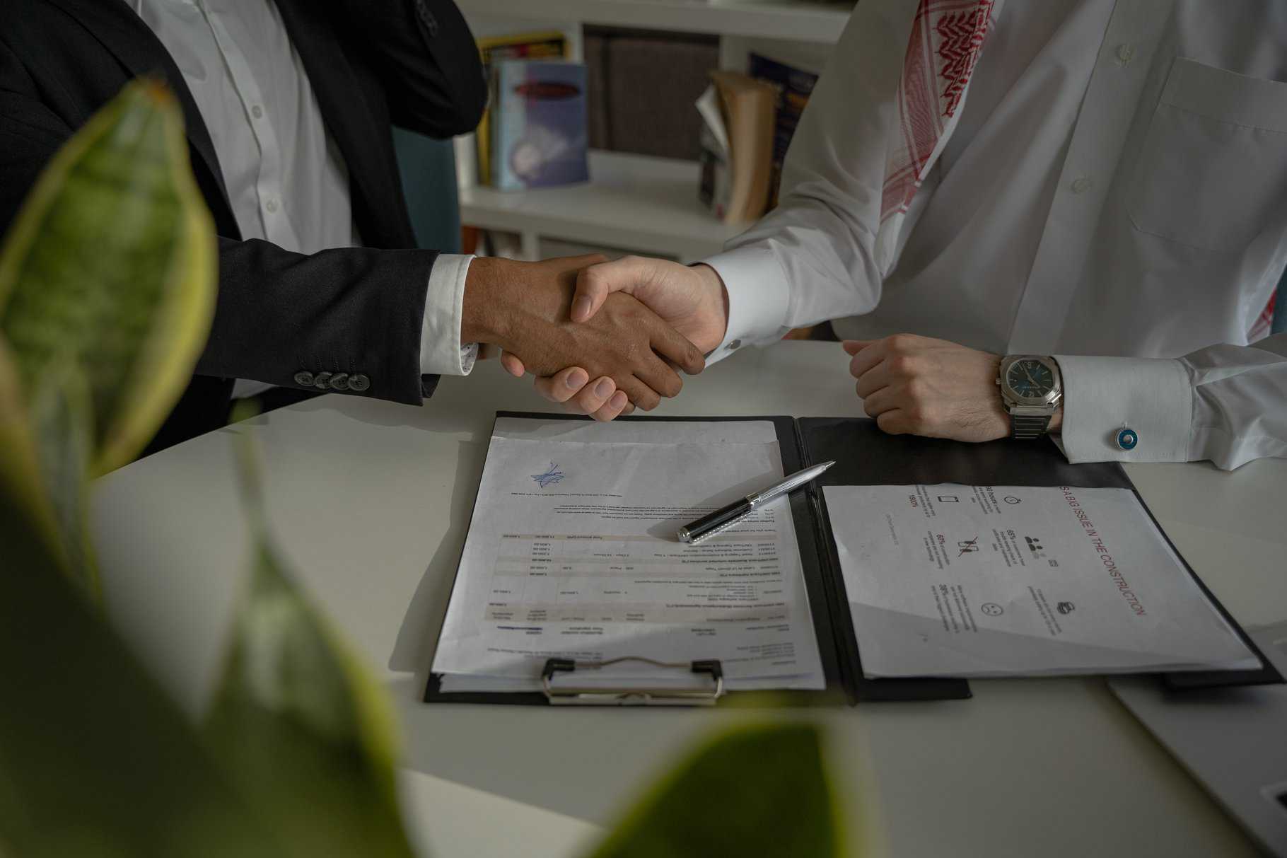 Two people are shaking hands over a table with documents and a pen on a clipboard, solidifying an agreement to resolve their property line dispute amicably.