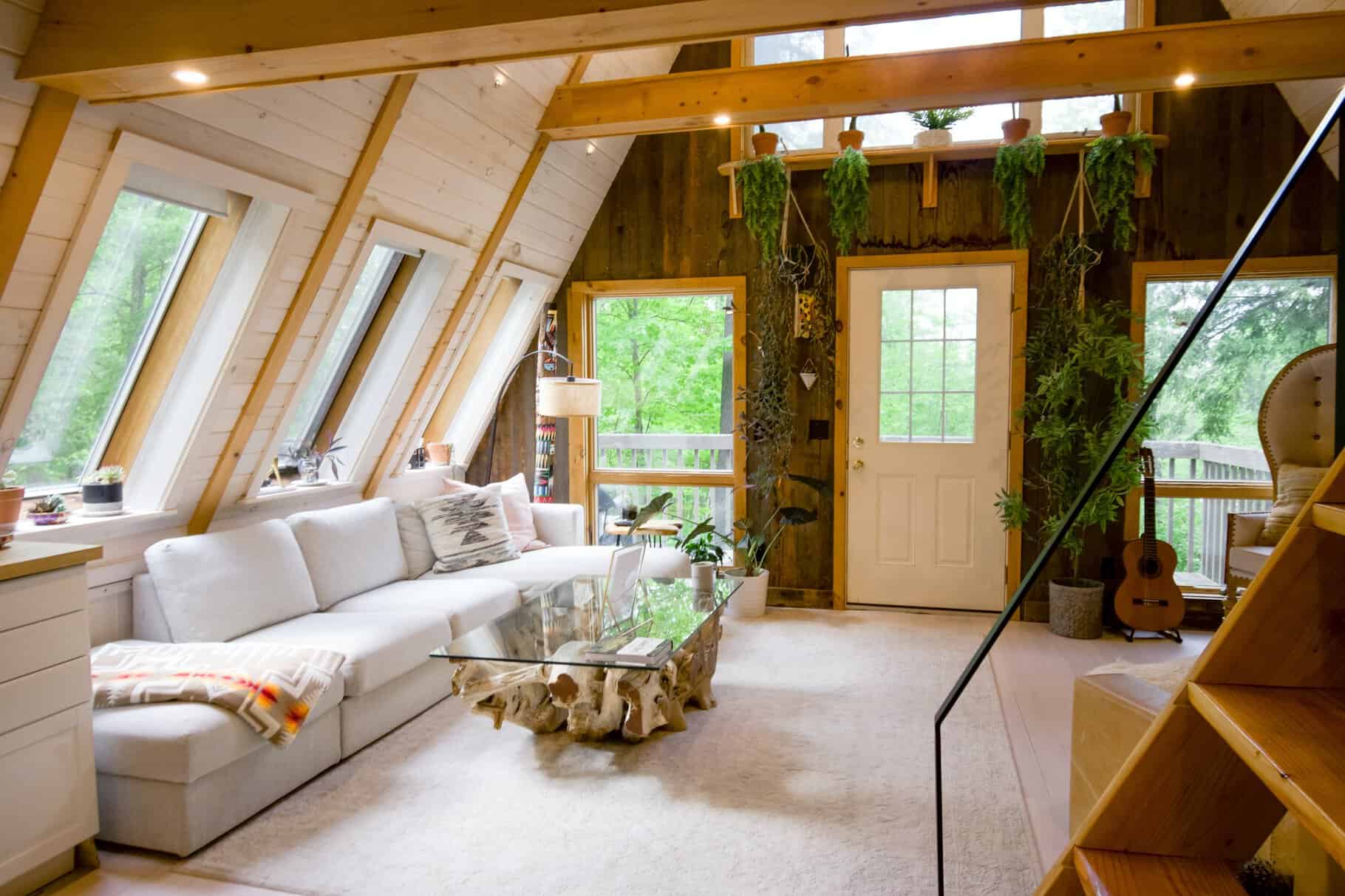 A-frame cabin interior with large windows, white sofa, glass coffee table, and plants hanging on wooden wall. Stairs lead to loft. Guitar and lamp in the corner. Light wood and greenery visible.