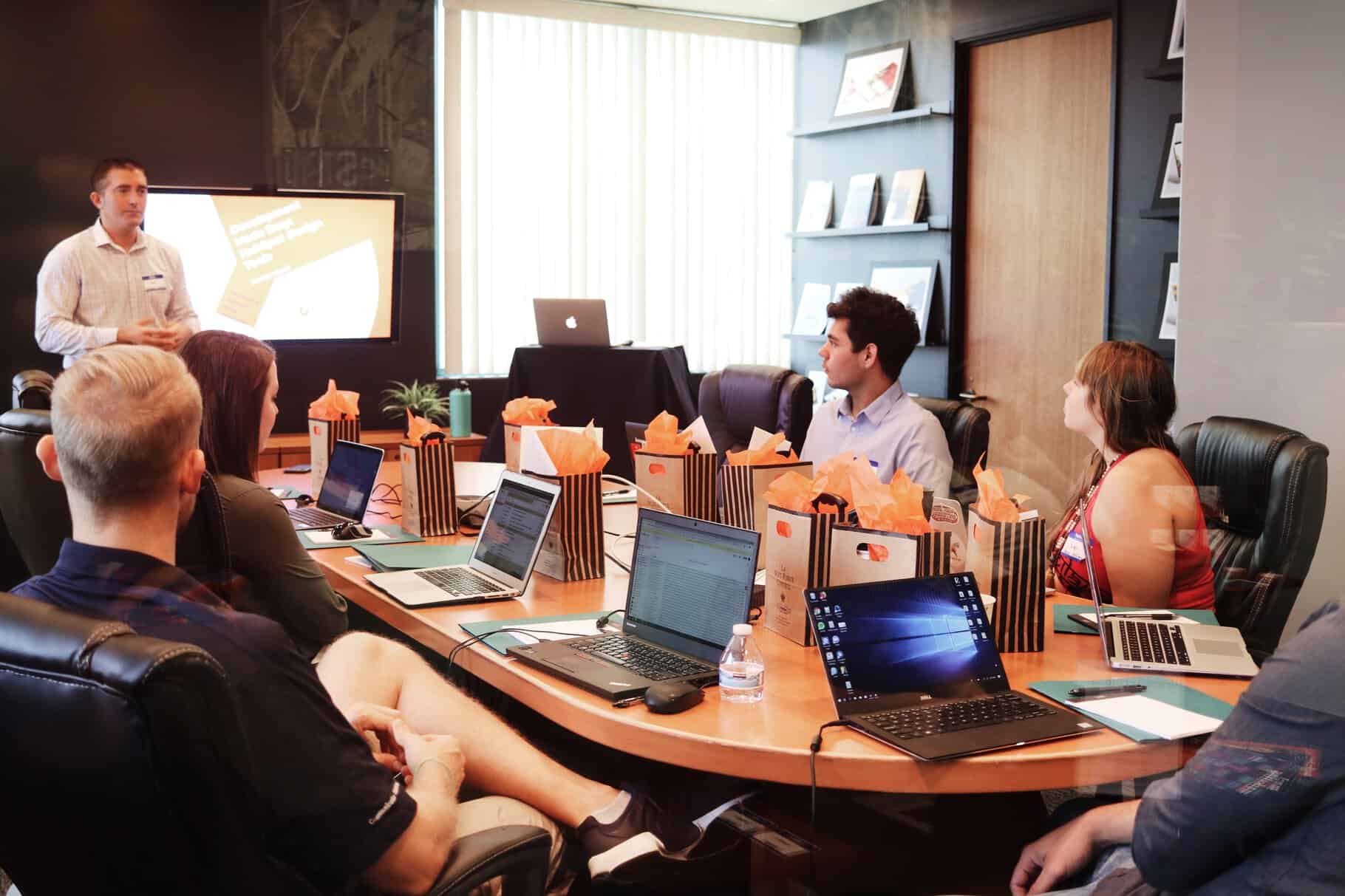 A group of people in a conference room are viewing a presentation. Laptops are open on the table, and small gift bags are set in front of each person.