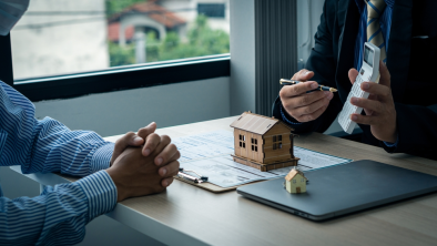 Two people discuss a real estate deal at a table with house models, documents, and a laptop. Amidst the negotiations, one person uses a calculator to clarify figures, while addressing concerns about potential property line disputes that could affect the agreement.