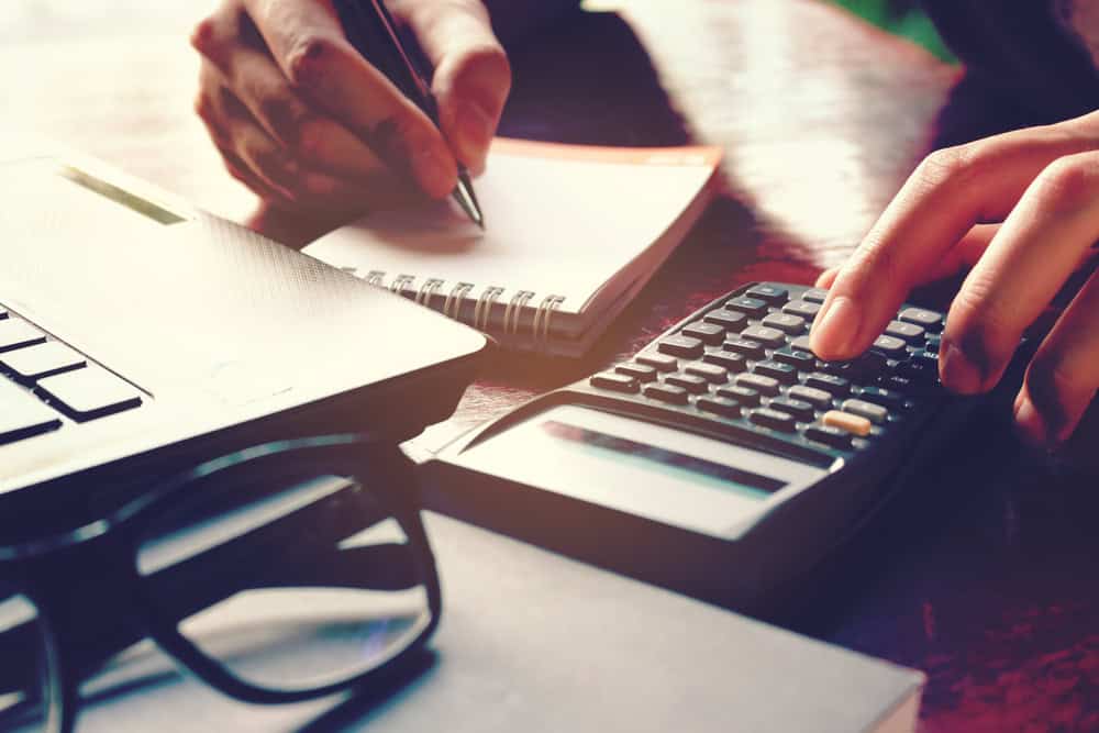 Hands using a calculator and jotting down notes in a notebook beside a laptop and glasses on a wooden table, meticulously calculating the finer points related to property line disputes.