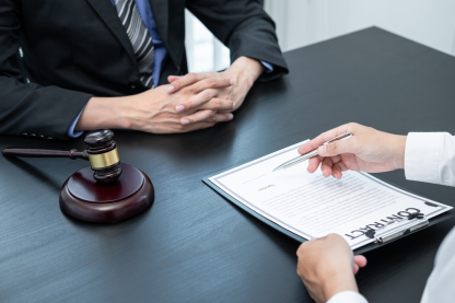 Two people in formal attire are seated at a table with a contract, gavel, and pen. One person points at the contract while the other listens attentively, discussing vital details of property line disputes.