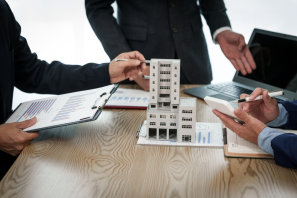 In a business meeting, three people engage in a discussion around a model building on the table, surrounded by laptops and documents with charts. The air is tense as they delve into concerns about property line disputes and their potential impact on the project's future steps.