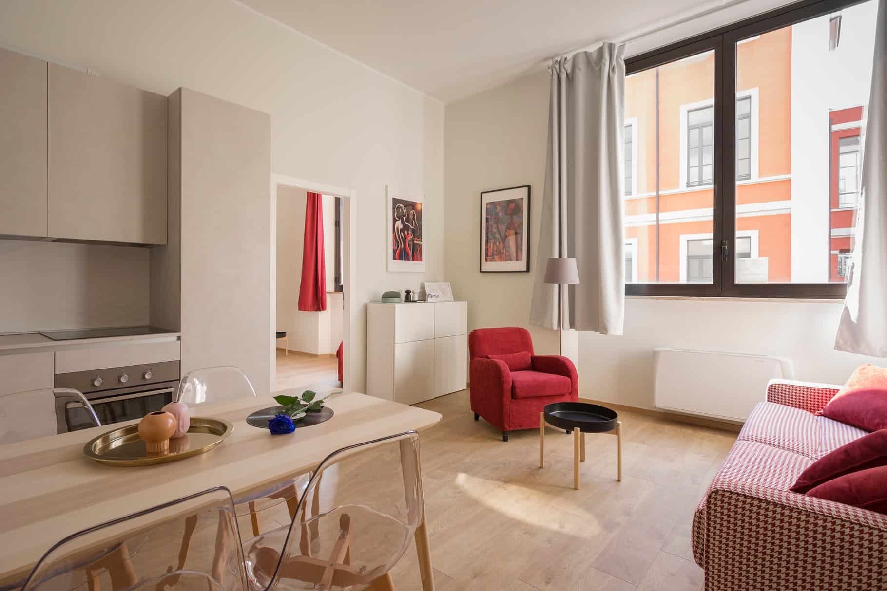 Modern living room with light wood flooring, featuring a red armchair, red sofa, small black table, and wall art. Large window lets in natural light. Kitchen and dining area with clear chairs.
