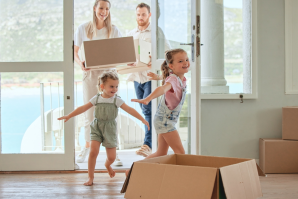 Two adults carry boxes into a room with large windows, while two children play near an open cardboard box, blissfully unaware of any property line disputes outside.
