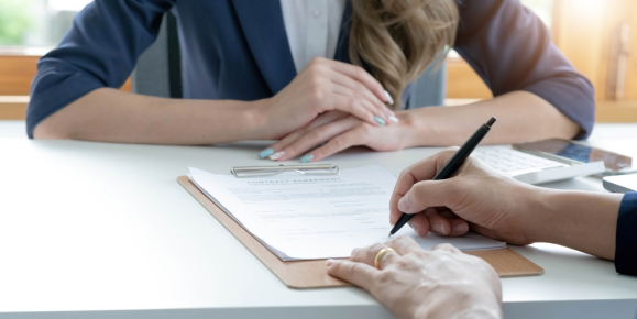 Two individuals sit at a desk; one signs a document on a clipboard while the other waits with hands clasped. A calculator rests in the background, silently hinting at the complexities of property line disputes being carefully navigated.