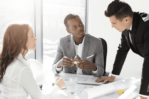 Three professionals in formal attire discuss architectural plans at a bright office table, navigating potential property line disputes. One holds a small house model, highlighting key boundary considerations.