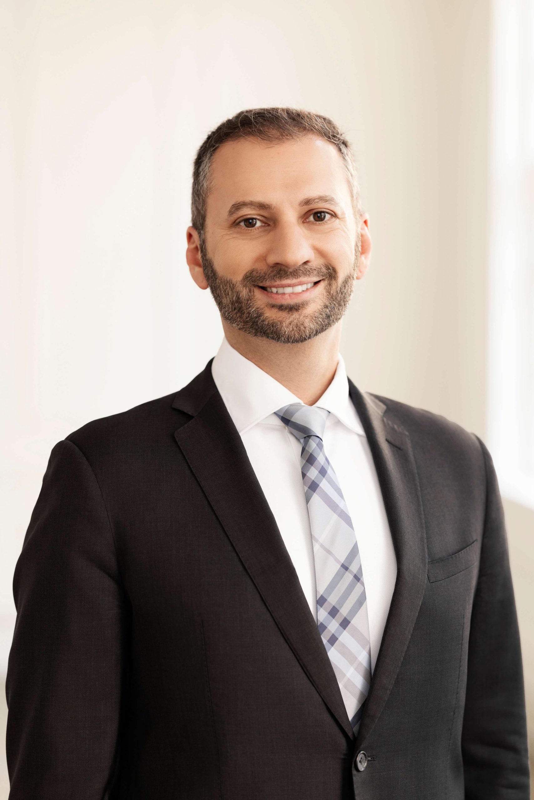 A man with short hair and a beard, wearing a dark suit, white shirt, and plaid tie, stands smiling against a plain background.