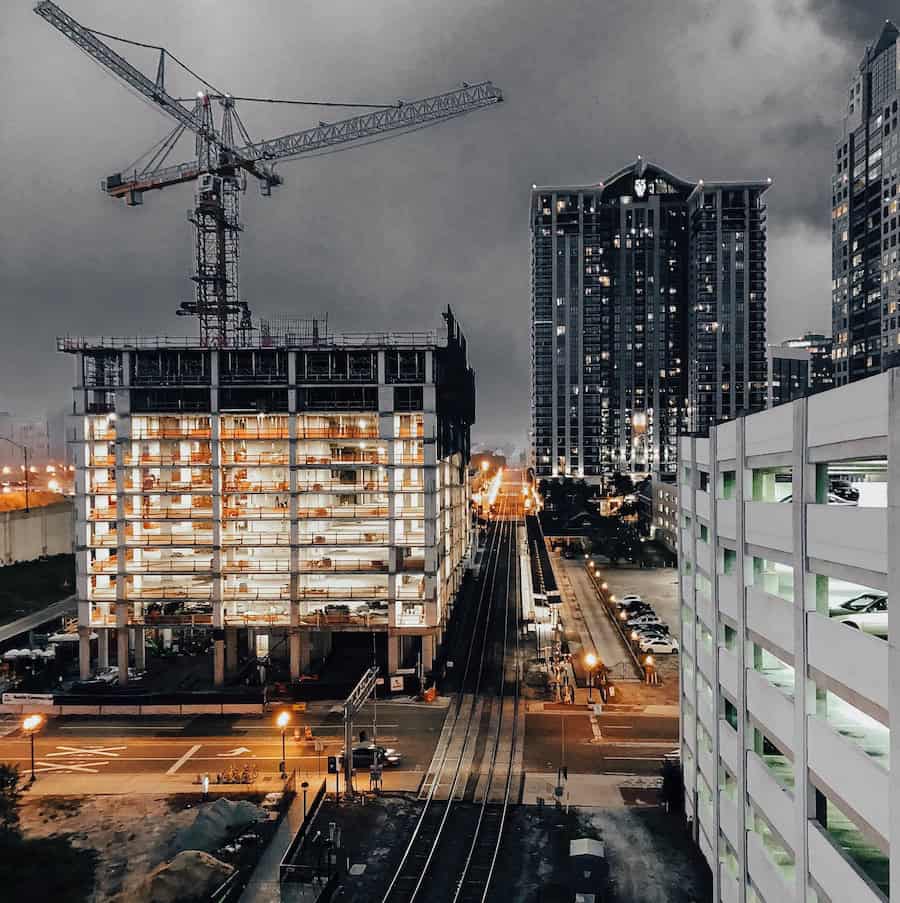 An urban nighttime scene unfolds with a lit high-rise construction site, where a crane hovers overhead. Adjacent tall buildings glimmer beside illuminated streets and train tracks below, all set under a cloudy sky, mirroring the complexities of property line disputes in the city's dynamic landscape.