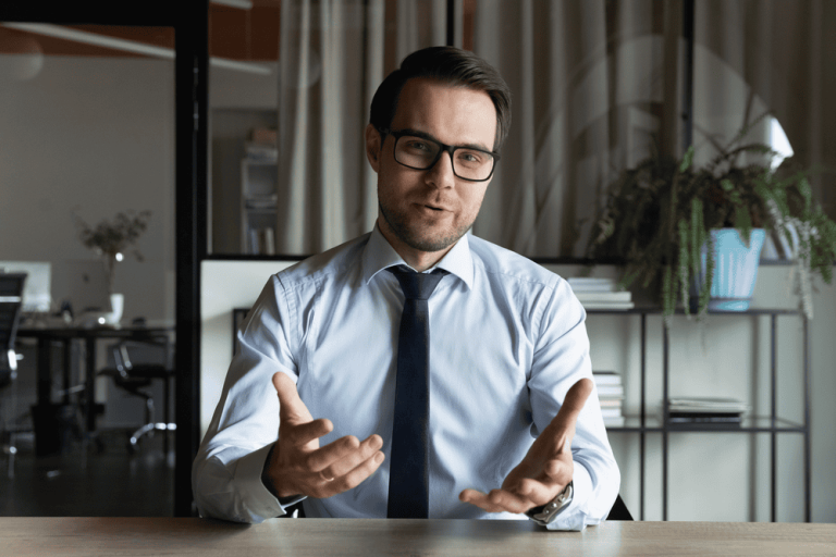 Man in glasses and a tie sits at a desk, gesturing with hands about property line disputes during a conversation in a modern office setting.