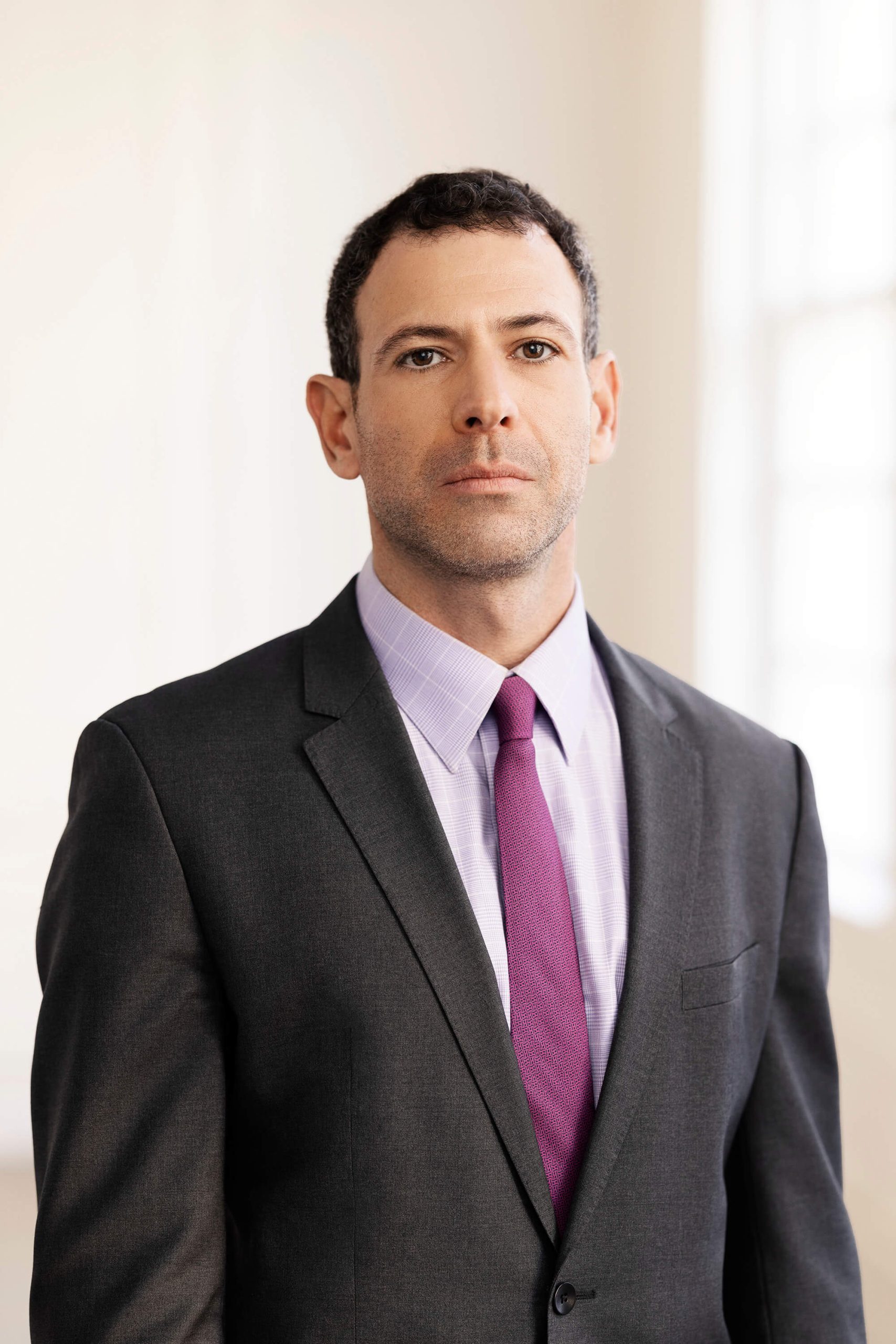 A man in a suit with a purple tie stands in a well-lit room, looking ahead with a neutral expression.