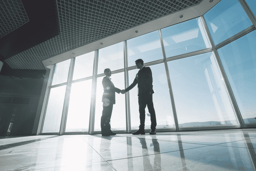 In a modern, sunlit room with large windows, two people in suits confidently shake hands, having just navigated the complexities of property line disputes with professionalism and mutual respect.