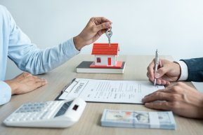 Two people sit at a table featuring a model house, contract, calculator, and money. One person signs the contract as the other holds a key, ready to settle any potential property line disputes with ease and assurance.