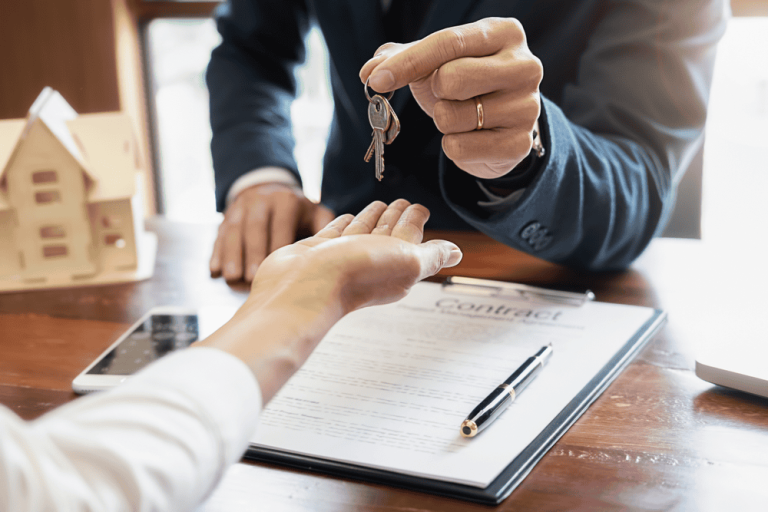 A person in a suit hands house keys to another across a table adorned with a contract and small house model, signifying not just the transfer of ownership but also the resolution of any looming property line disputes.