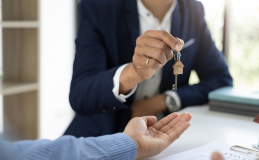 In a professional office setting, a person in a suit hands over a house-shaped keychain to another, symbolizing the resolution of property line disputes with mutual understanding and agreement.