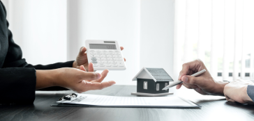 Two people discuss home financing. One hand holds a calculator, the other points to a miniature house model on a desk filled with documents. As they talk numbers, property line disputes emerge as an unexpected factor needing careful consideration in their planning.
