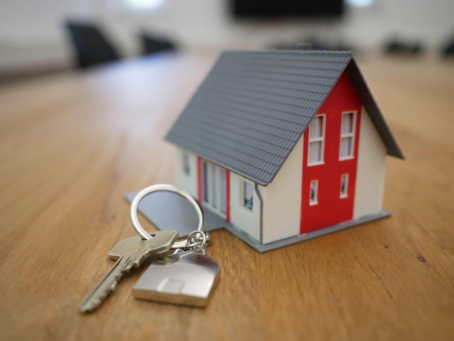 A small model house and a set of keys with a house-shaped keychain are placed on a wooden table.