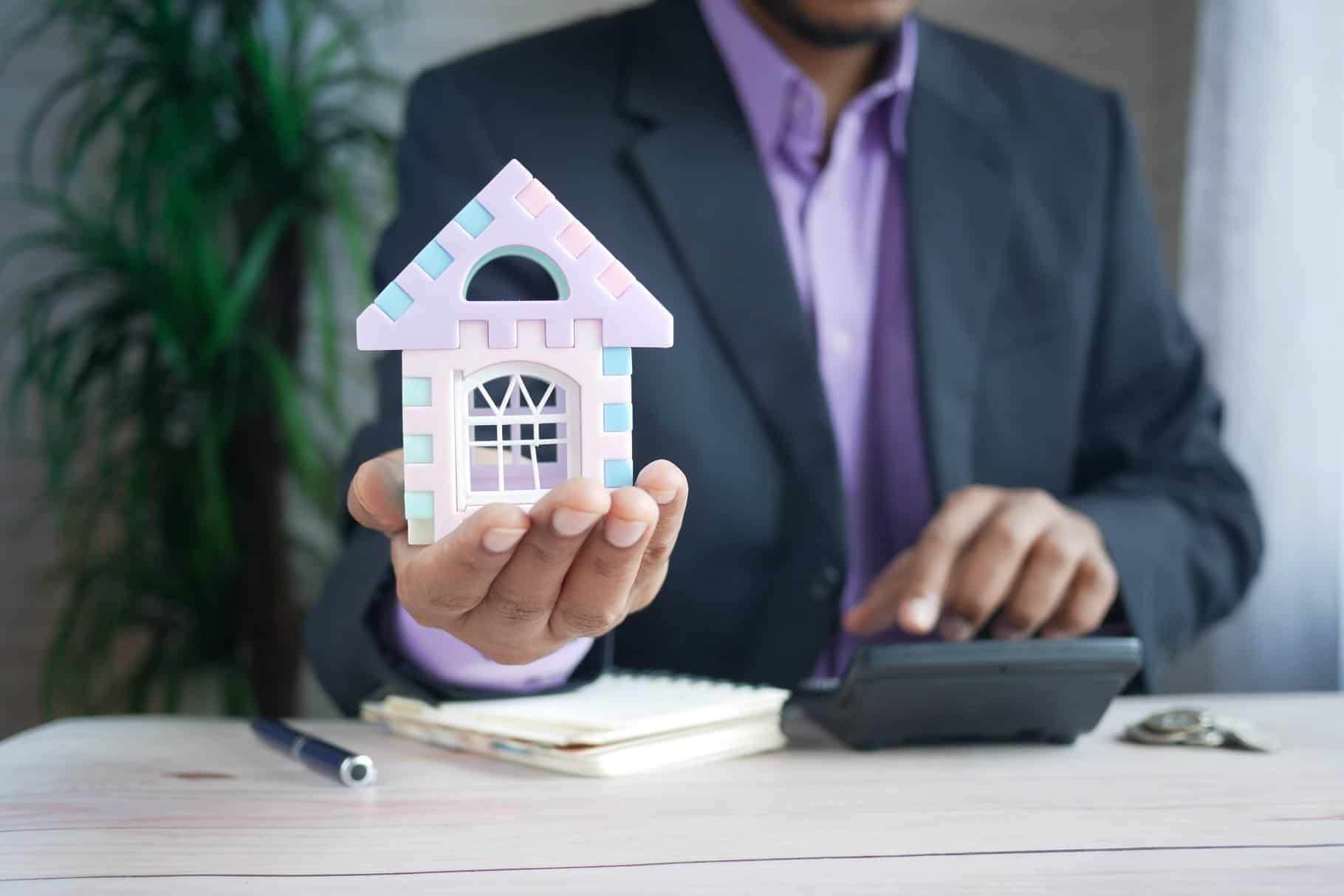 A person in a suit holds a colorful toy house model, with a pen, notebook, and calculator on the desk.