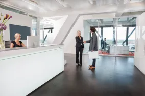 A modern office lobby features a receptionist at the desk. In the background, two individuals—one clutching documents—are shaking hands near a glass partition, likely discussing property line disputes. Another person is visible through the glass, hinting at more consultations to come.