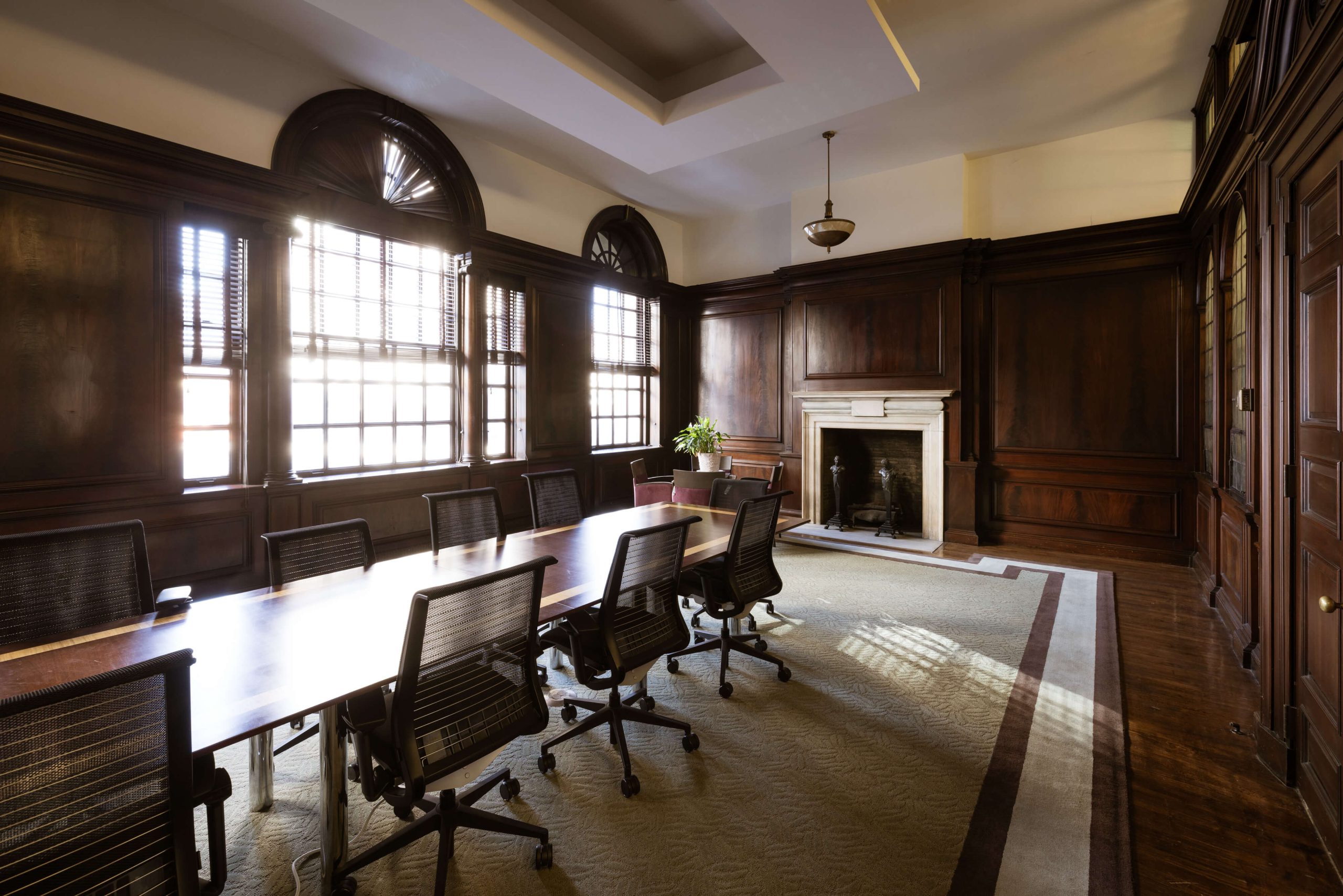 A conference room with a long wooden table, office chairs, large windows, wood-paneled walls, a fireplace, and a potted plant.