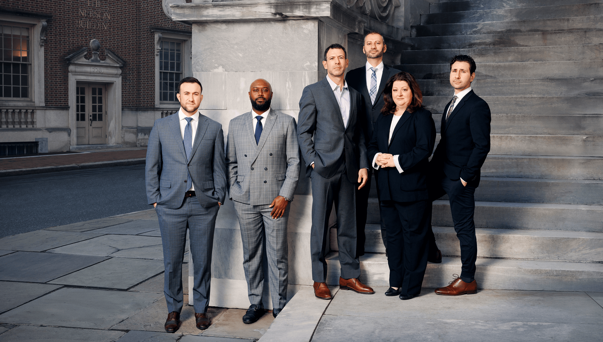 Six people in business attire stand on a stone walkway and steps outside a large building, posing for a formal group photo.