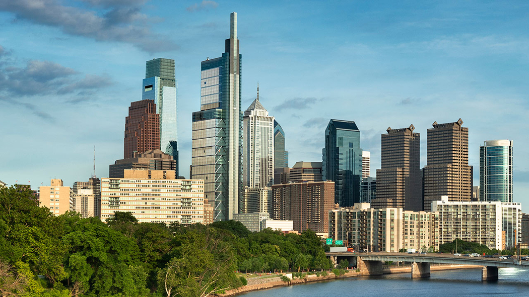 Philadelphia skyline with modern and historic high-rise buildings under a blue sky, trees in the foreground, and a river and bridge on the right.