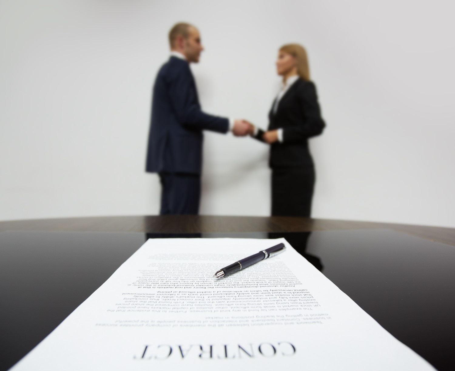 A pen and contract are on a table in the foreground, while a man and woman in business attire shake hands in the blurred background—perfect for illustrating a meeting with a Non-compete Agreement Lawyer.