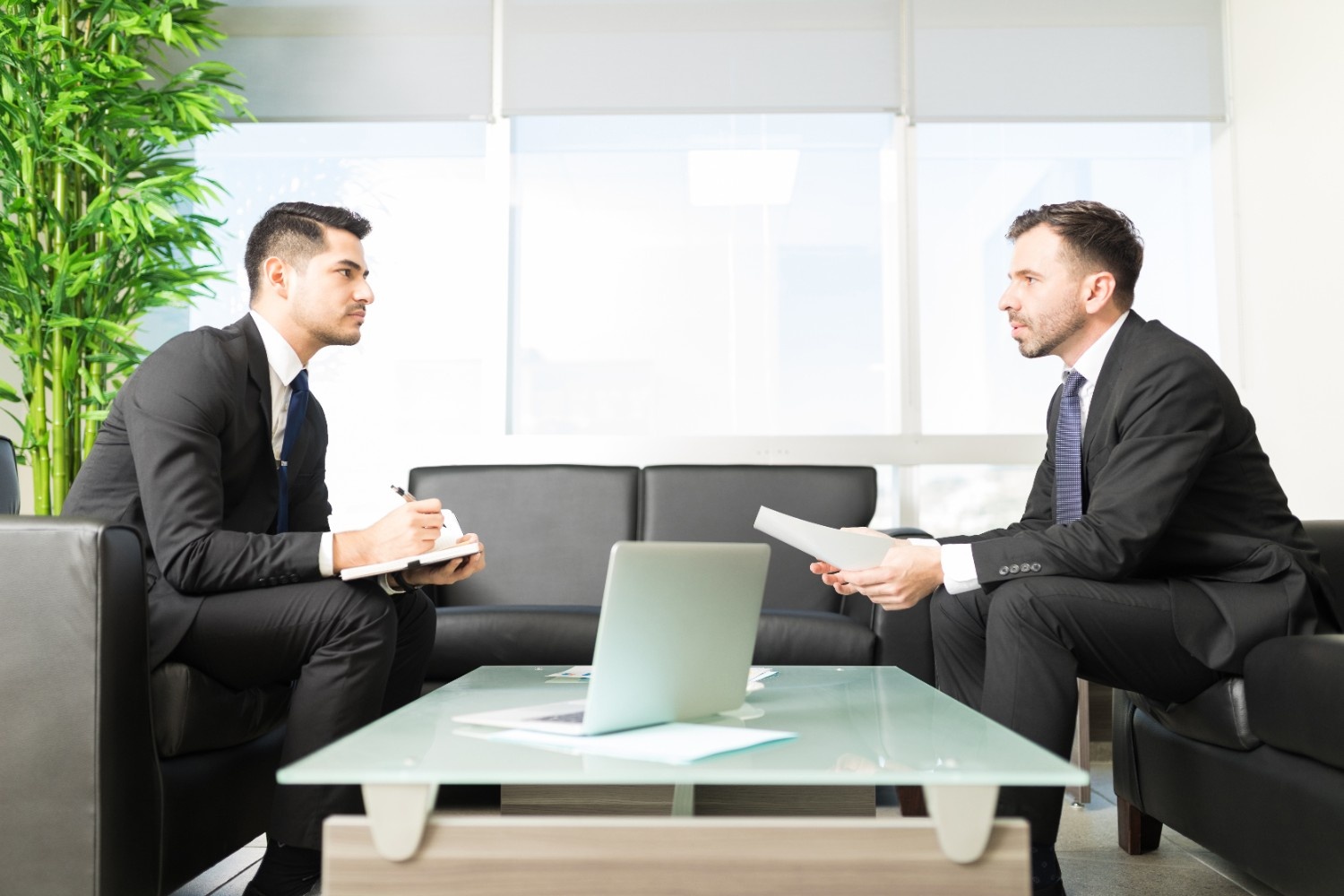 Two men in business attire sit across from each other in an office, discussing documents and consulting a laptop as they review matters with their Outside General Counsel.