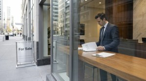suited professional reviewing documents near For Sale sign outside urban building
